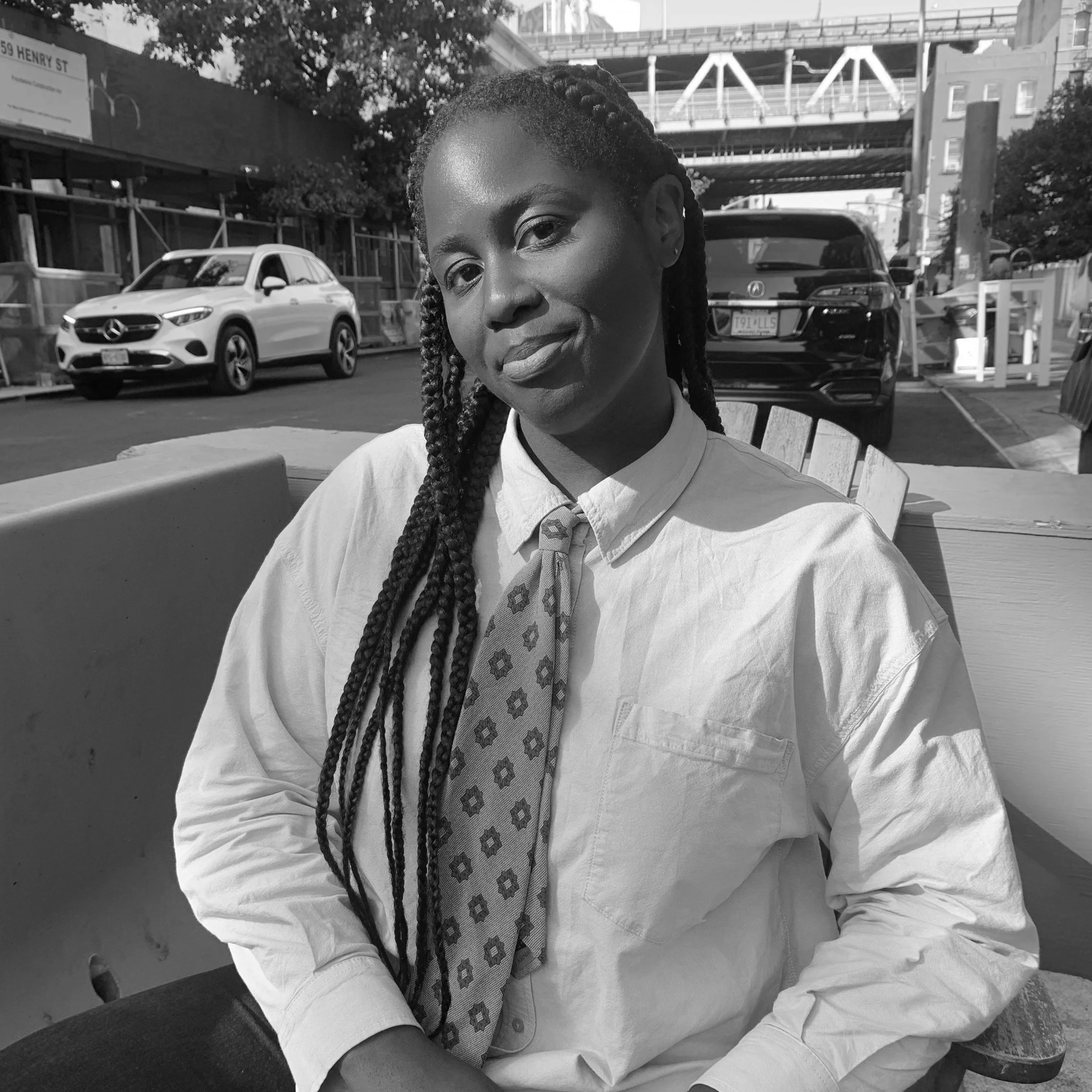 A woman with braided hair smiles while sitting outdoors at a table, wearing a white shirt and a patterned tie, with cars and buildings visible in the background.