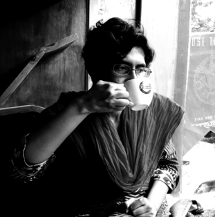 A woman with short curly hair and glasses drinking from a cup inside a coffee shop