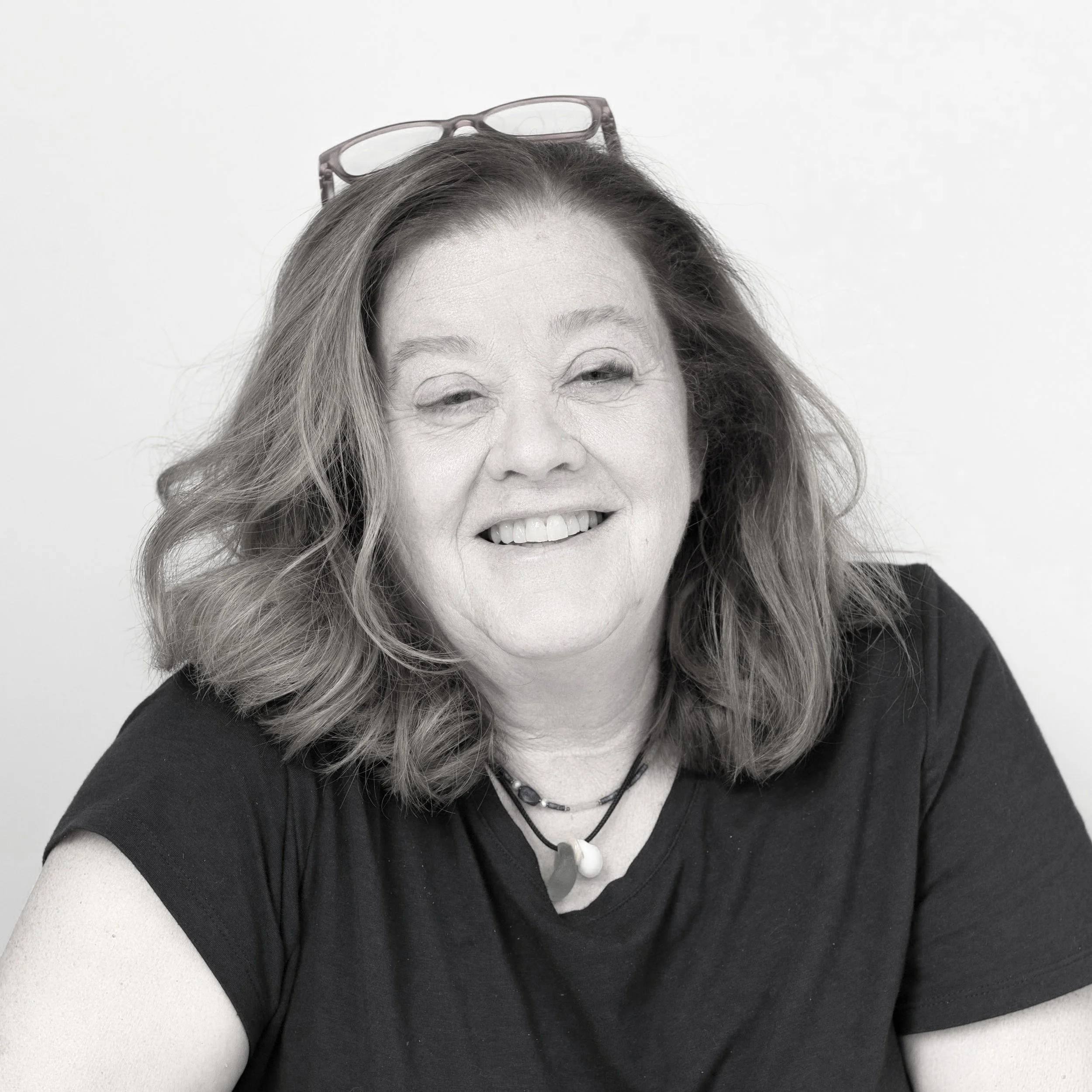 Black and white portrait of a woman with wavy hair, wearing glasses on her head, smiling, and wearing a black shirt with a necklace.