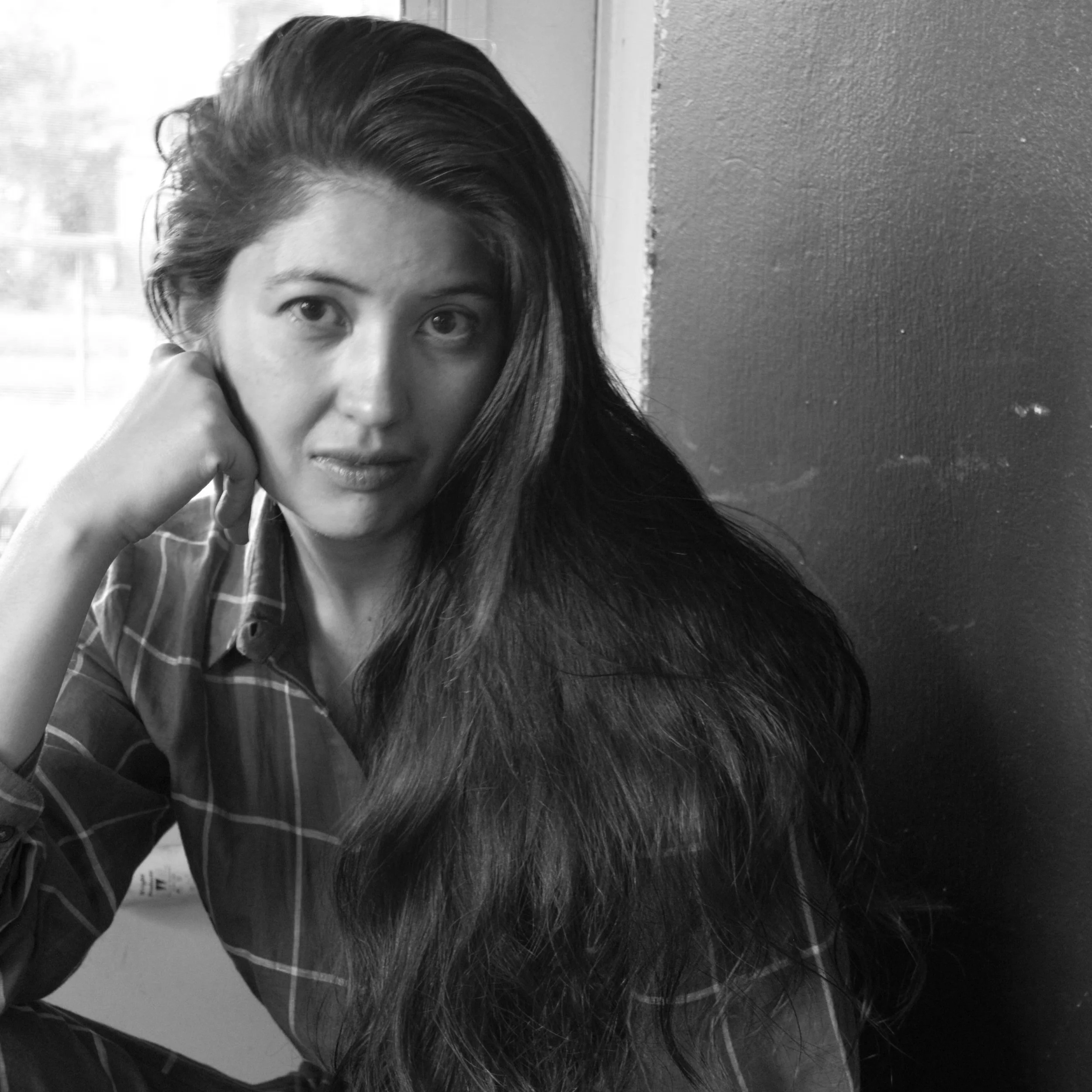 Black and white photo of a woman with long dark hair, wearing a plaid shirt, resting her head on her hand, sitting near a window.