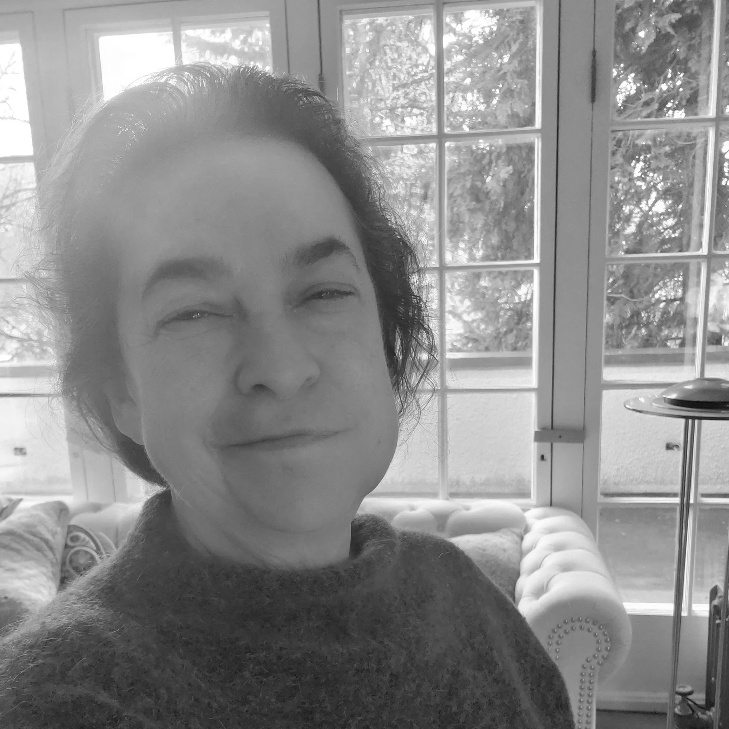 A woman with short dark hair making a facial expression with squinted eyes and a slight smile, standing in a room with large windows and a white tufted sofa in the background.