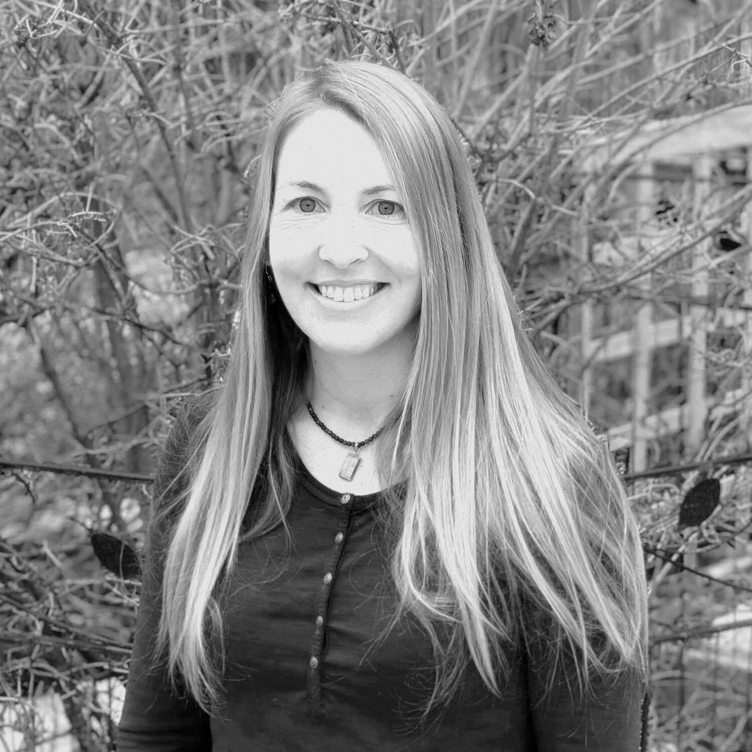 Black-and-white portrait of a woman with long, straight hair, smiling, standing outdoors in front of leafless bushes or trees.
