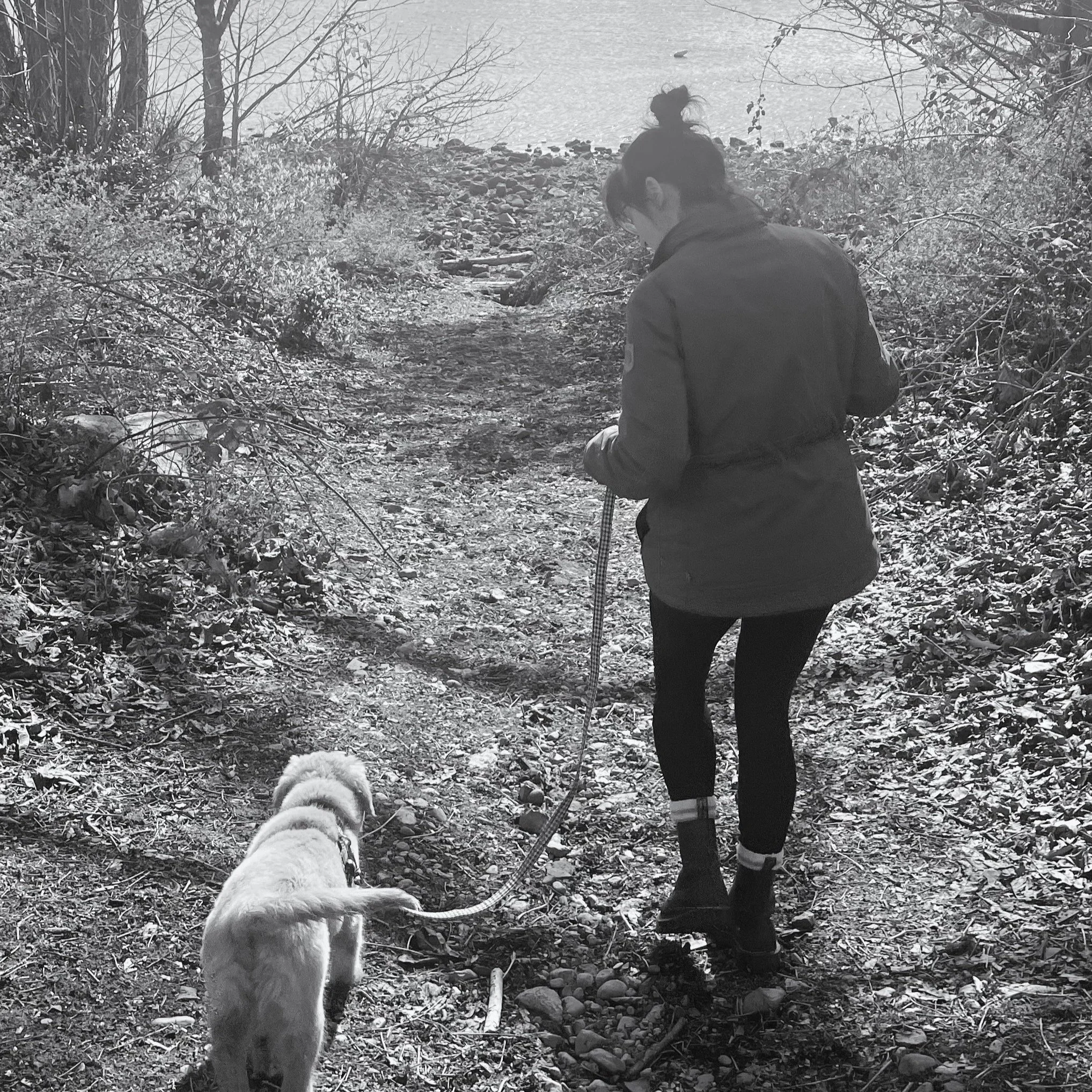 A woman walking her dog on a trail near a body of water, wearing a coat and boots.
