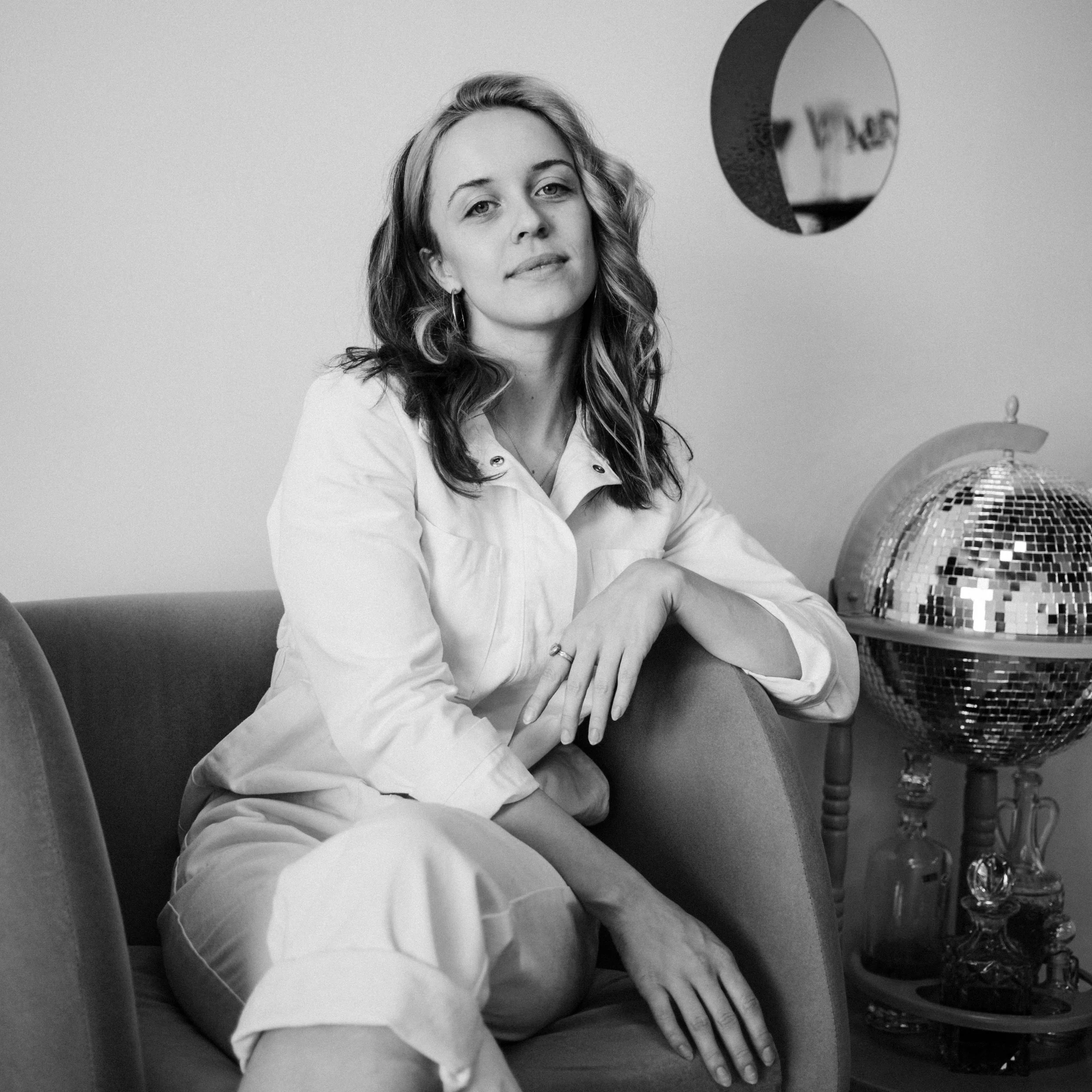 Black and white photo of a young woman with wavy hair sitting on a sofa, wearing a light-colored shirt, with a relaxed pose and confident expression. There is a disco ball and a shelf with bottles in the background.