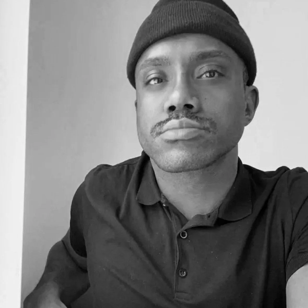 Black and white photo of a man wearing a beanie and a collared shirt, looking at the camera with a neutral expression.