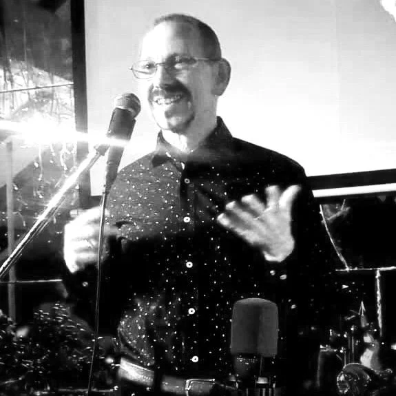 A man with glasses and a goatee, smiling and speaking into a microphone, gesturing with his right hand, in a dimly lit room with a window and neon lights.