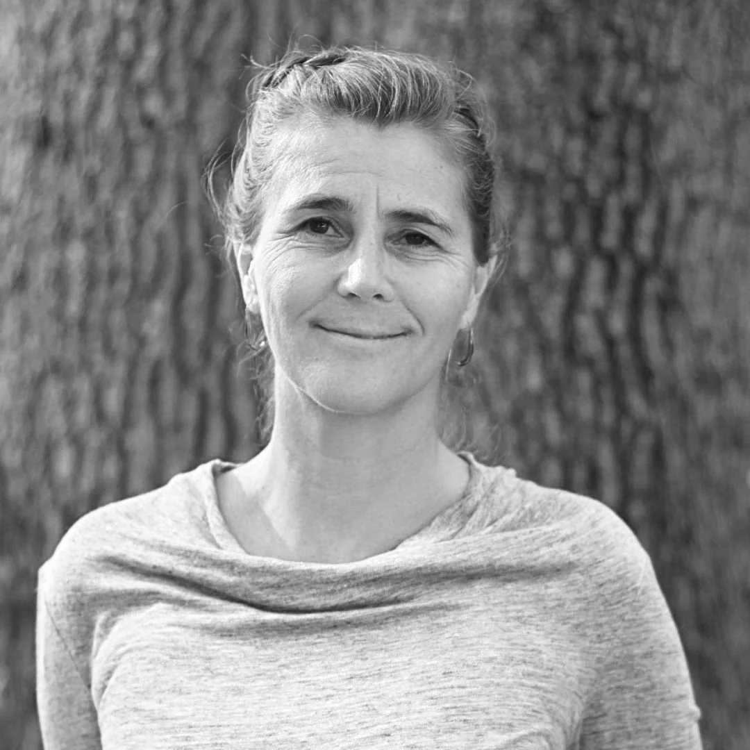Black and white portrait of a woman outdoors with a tree trunk in the background, wearing a light-colored shirt and looking at the camera with a gentle smile.