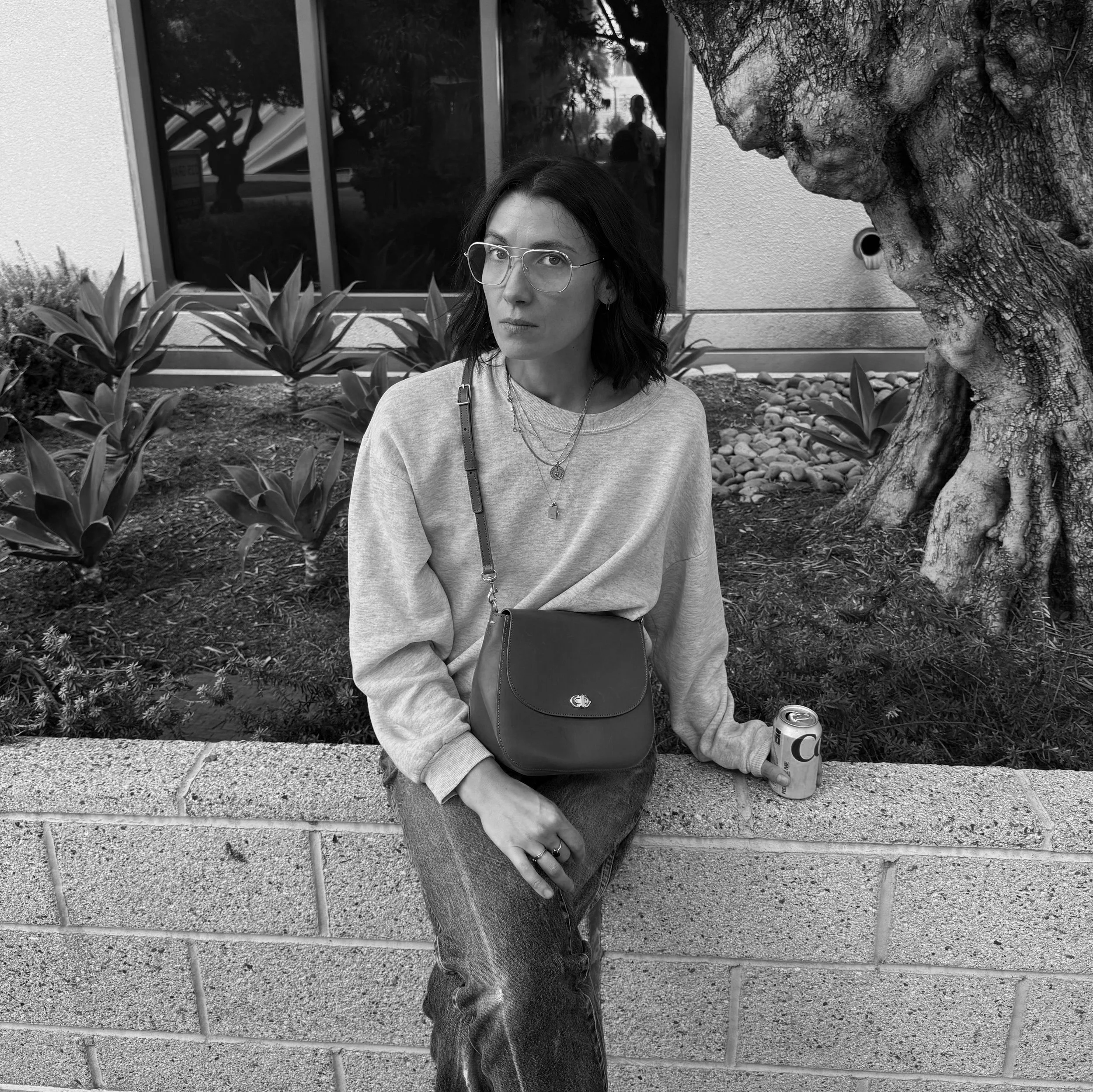 A woman with glasses and shoulder-length dark hair sitting on a brick wall, holding a can of soda in her right hand, with a garden and tree in the background.
