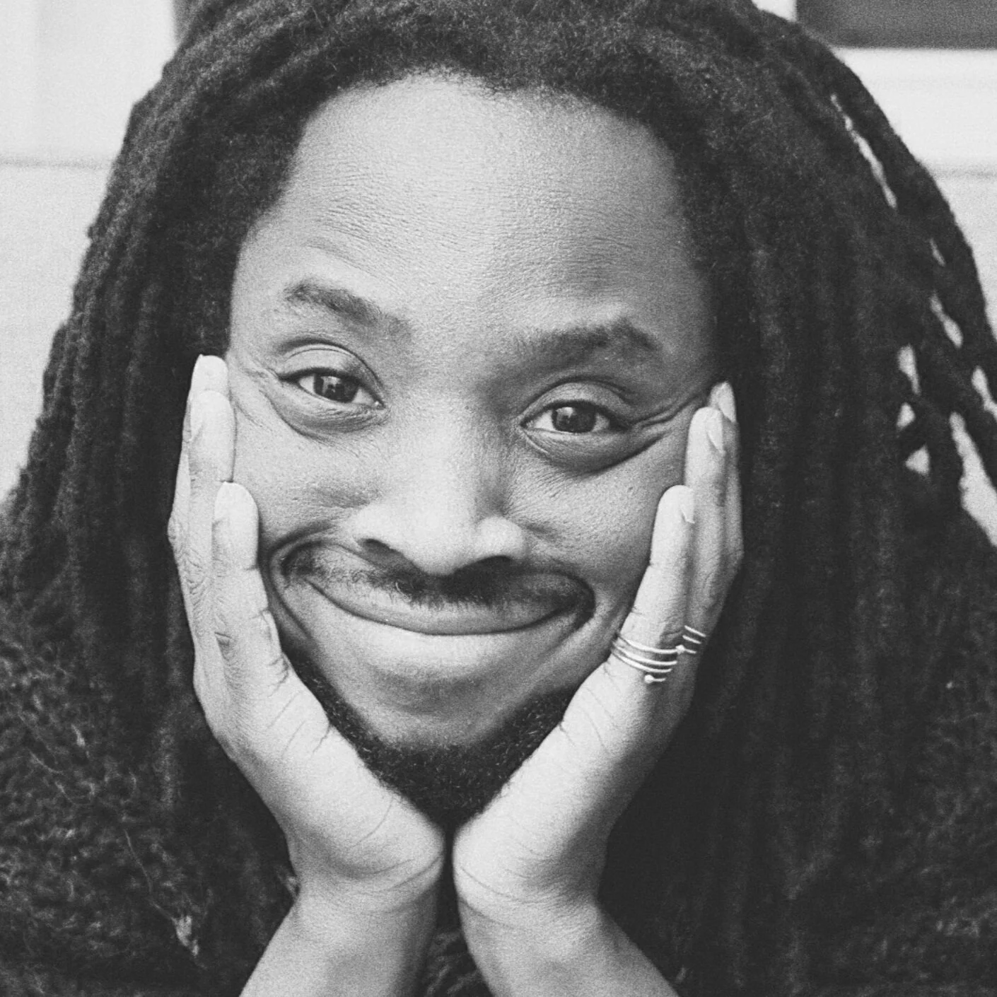 A black and white photo of a smiling person with dreadlocks, resting their face in their hands.