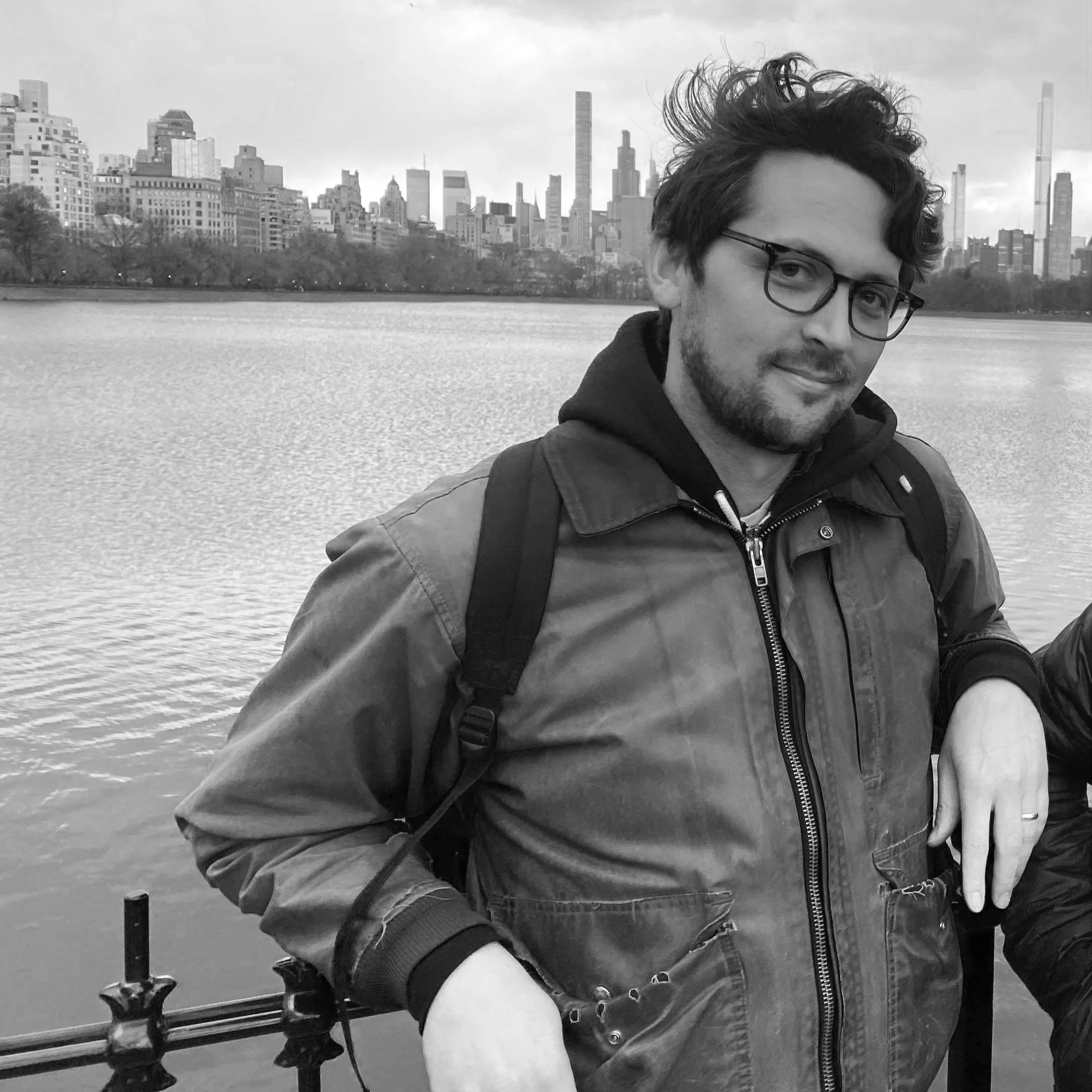 A man with glasses and a beard wearing a jacket and backpack, standing near a waterfront with a city skyline in the background.