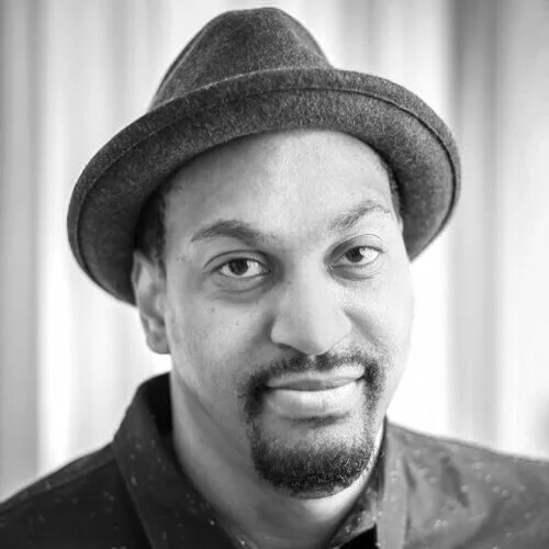 Black man wearing a fedora hat and a dark shirt, smiling at the camera.