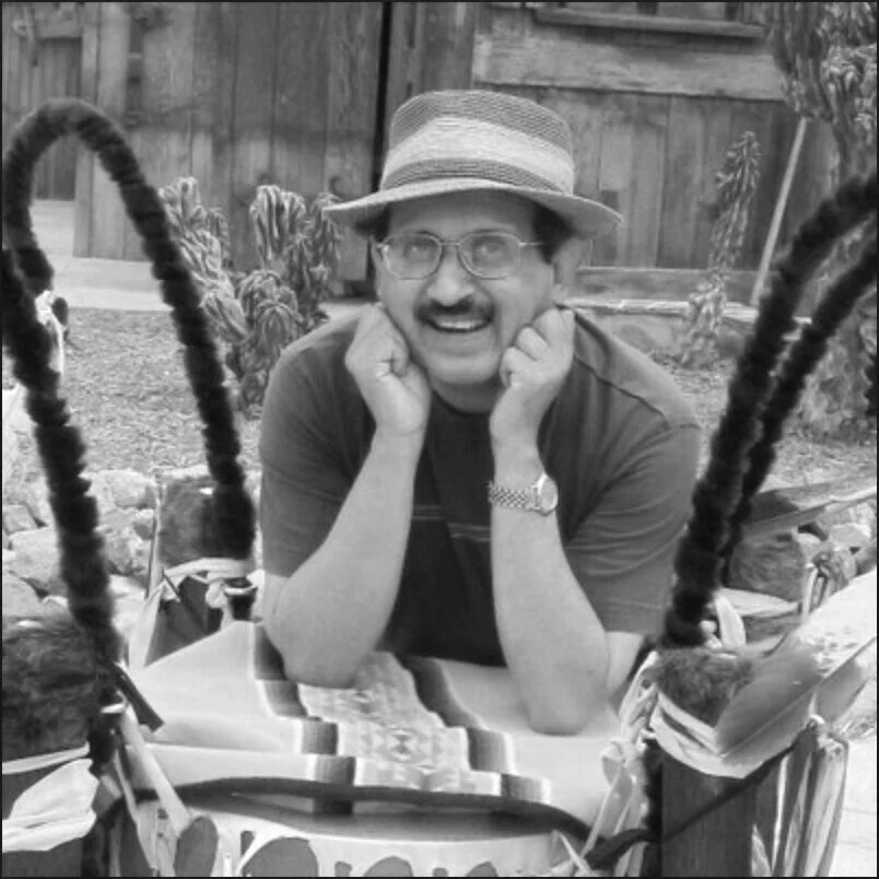 A man with glasses and a mustache smiling, wearing a straw hat, sitting outdoors at a table with a colorful cloth, with cacti and a wooden fence in the background.