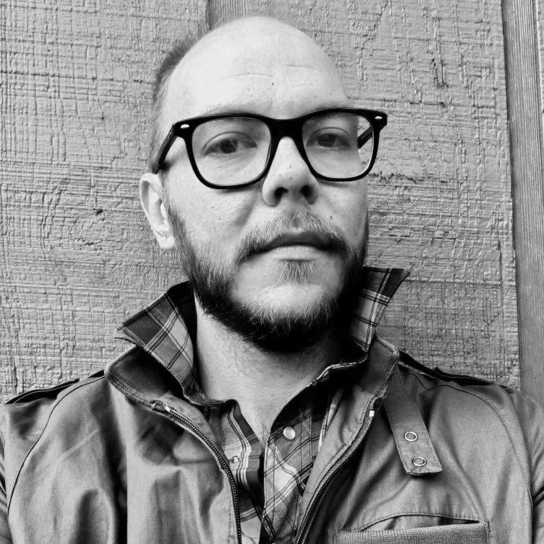 A man with glasses and a beard taking a selfie in front of a wooden wall.