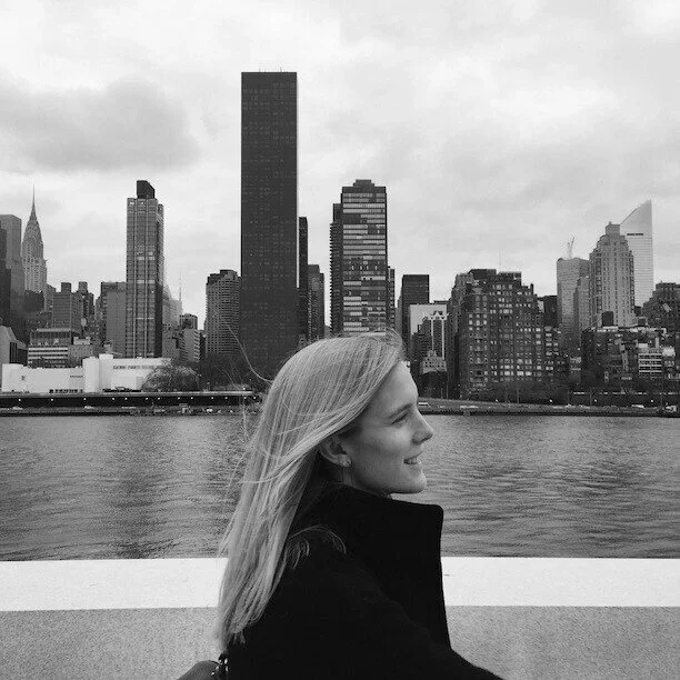 A woman with long blonde hair standing by a river with city skyscrapers in the background in New York City.