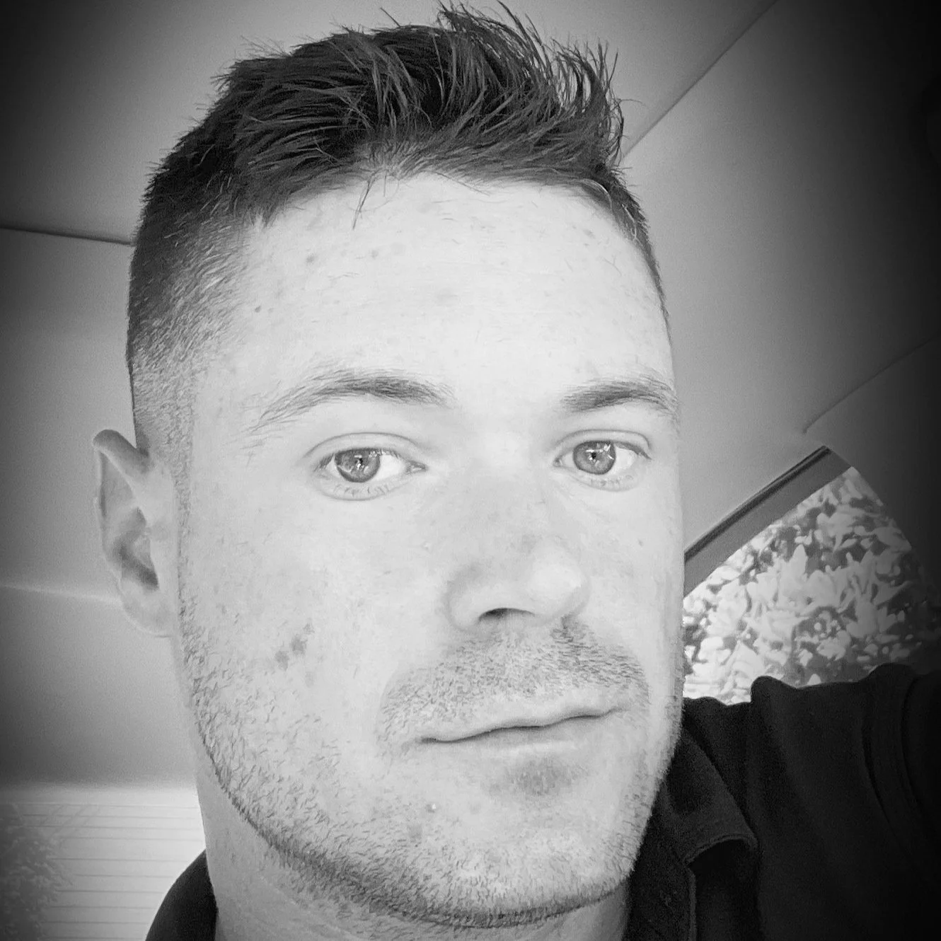 Close-up black and white photo of a young man with short dark hair, light eyes, and a slight stubble, looking directly at the camera with a neutral expression.