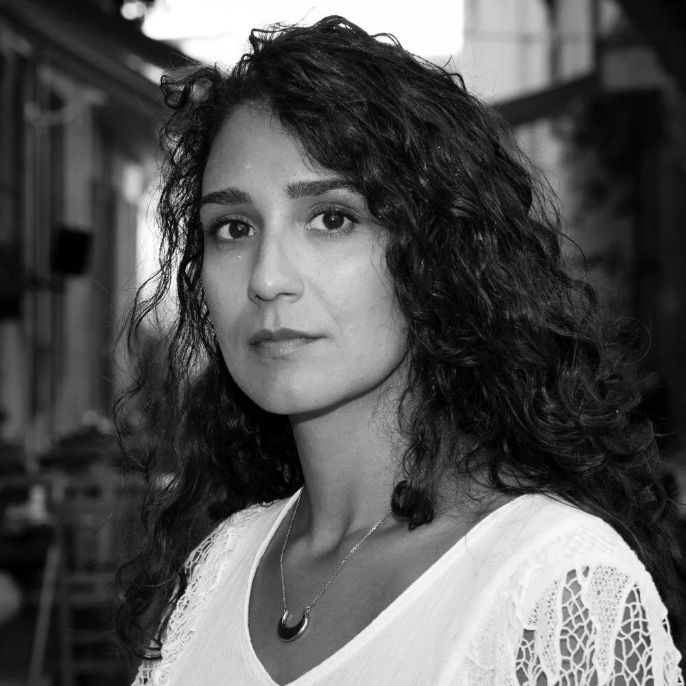 Black and white portrait of a woman with curly hair, wearing a white top with lace details and a necklace, outdoors setting.