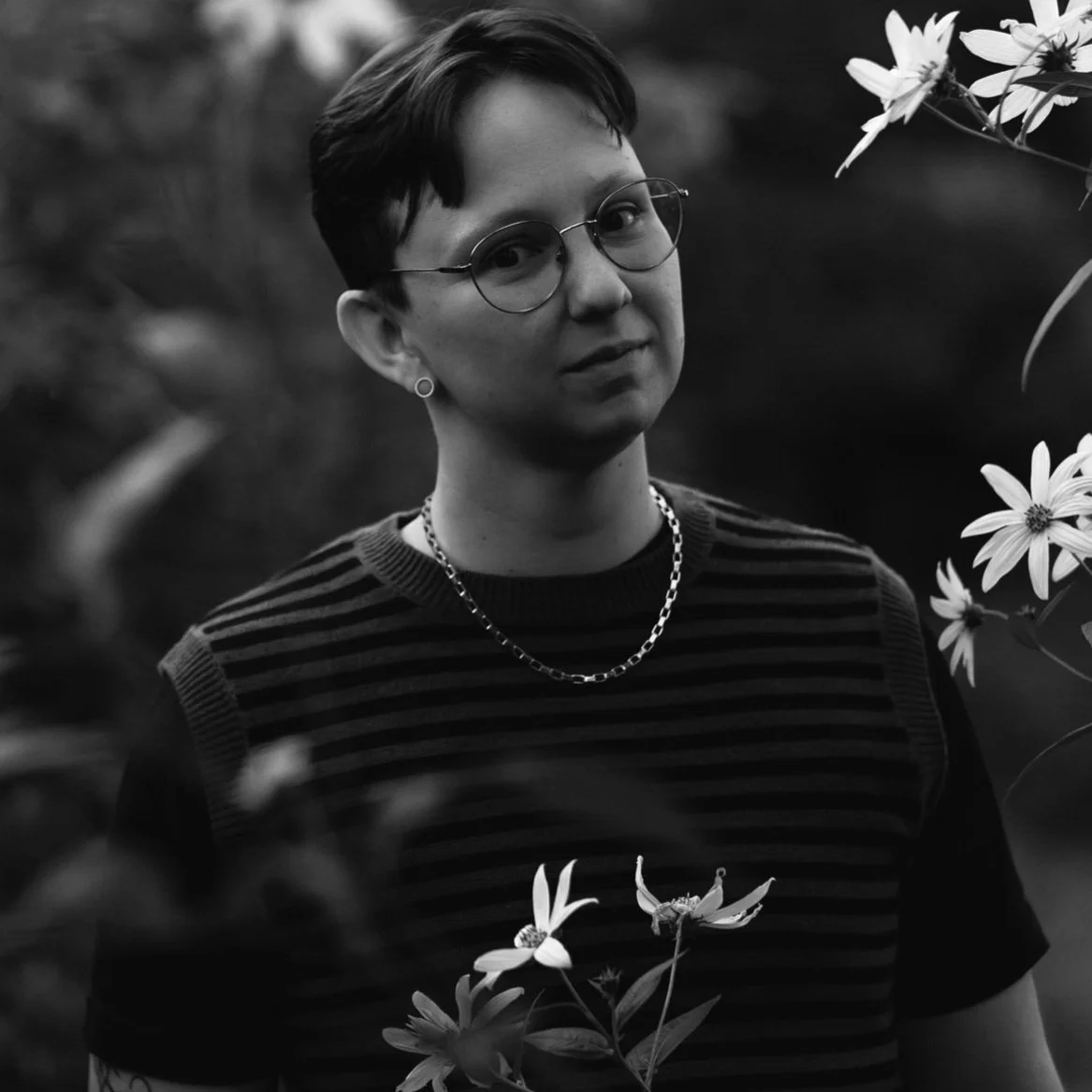 Black and white photo of a person with short hair, round glasses, wearing a striped shirt, necklace, and earrings, standing among flowers outdoors.