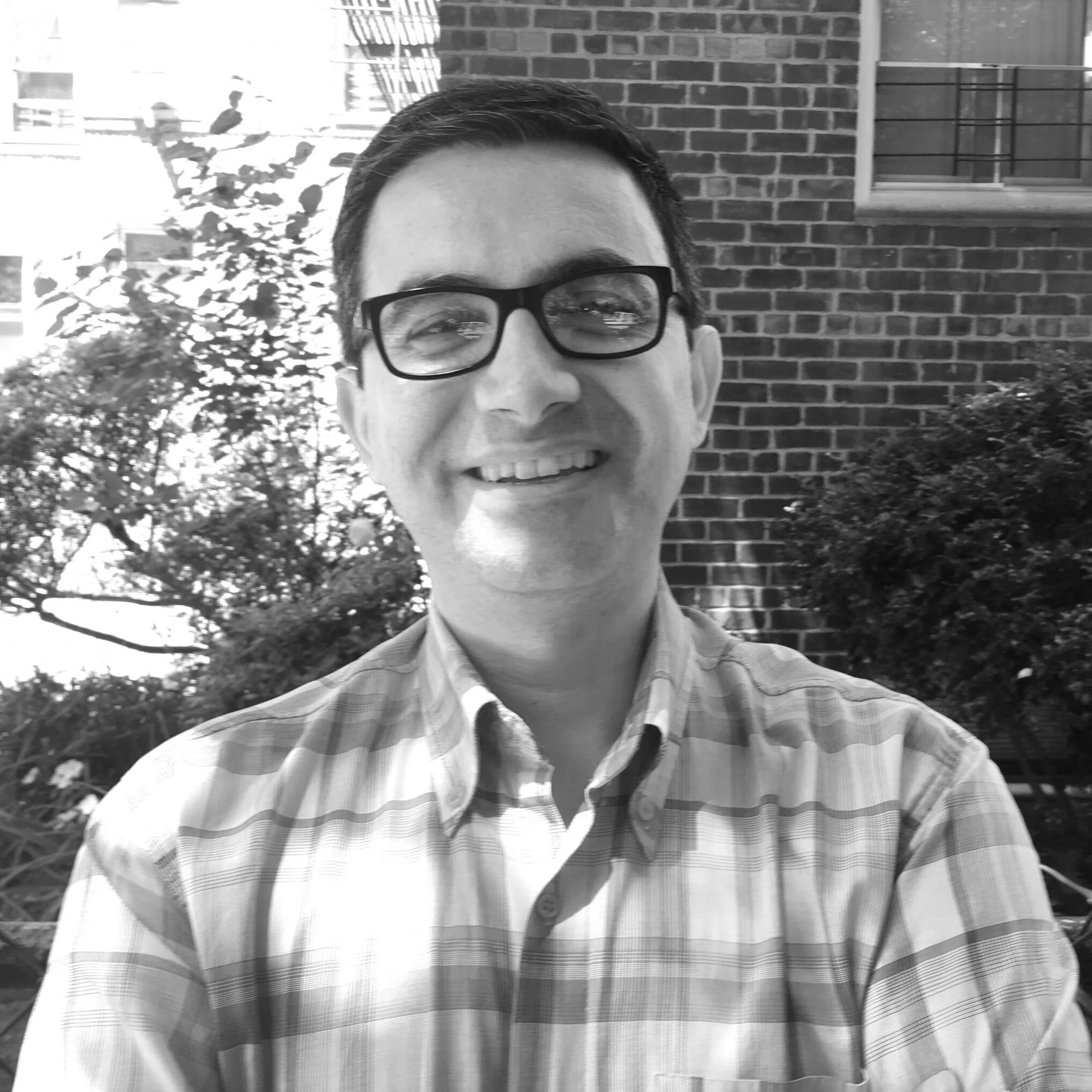 A smiling man with glasses and short dark hair taking a selfie outdoors in front of a brick building, bushes, and tree branches in black and white.
