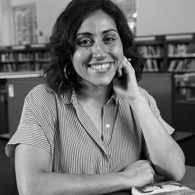 A woman with shoulder-length dark hair smiling, sitting at a table in a library, wearing a striped short-sleeve shirt, with bookshelves in the background.
