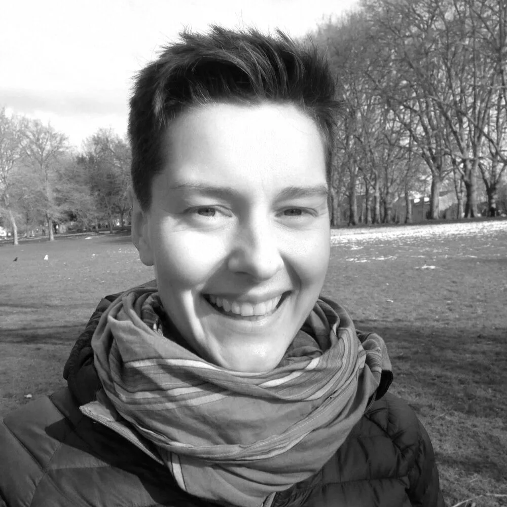 Young man smiling outdoors in a park with trees and open field in the background, wearing a scarf and jacket on a cloudy day.