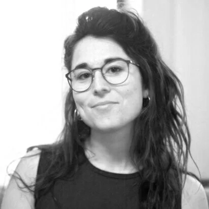 A young woman with wavy dark hair, wearing glasses and a sleeveless top, smiling slightly in a black-and-white photo.