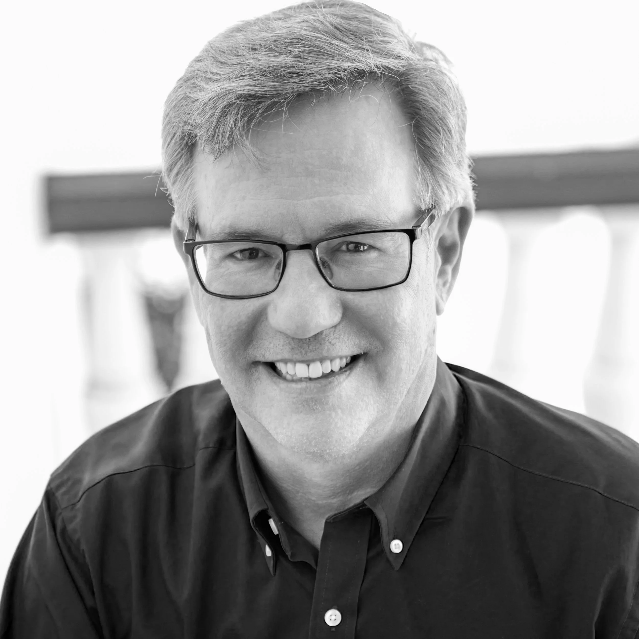 Black and white portrait of a smiling man with glasses, short light hair, wearing a dark collared shirt, with a blurred background.