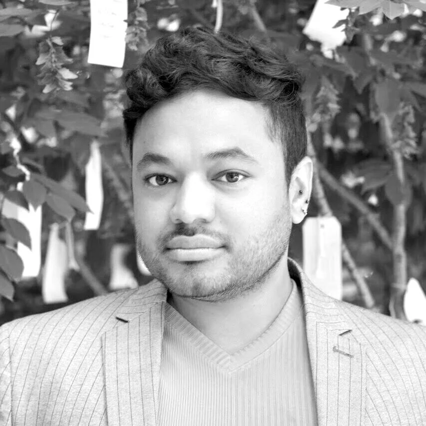 A black and white portrait of a young man with short, curly hair and earrings, wearing a striped blazer and shirt, standing in front of blurred foliage with tags hanging from the trees.