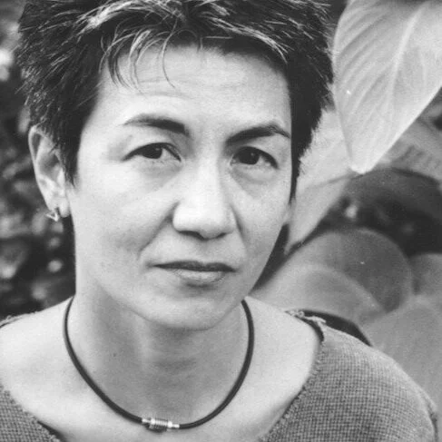 Black and white portrait of a woman with short hair, wearing a necklace and a sweater, with a plant in the background.