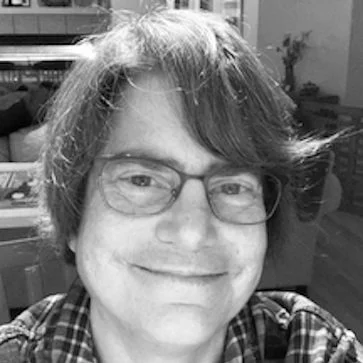 A smiling person with glasses and medium-length hair, indoors with household items and a potted plant in the background.