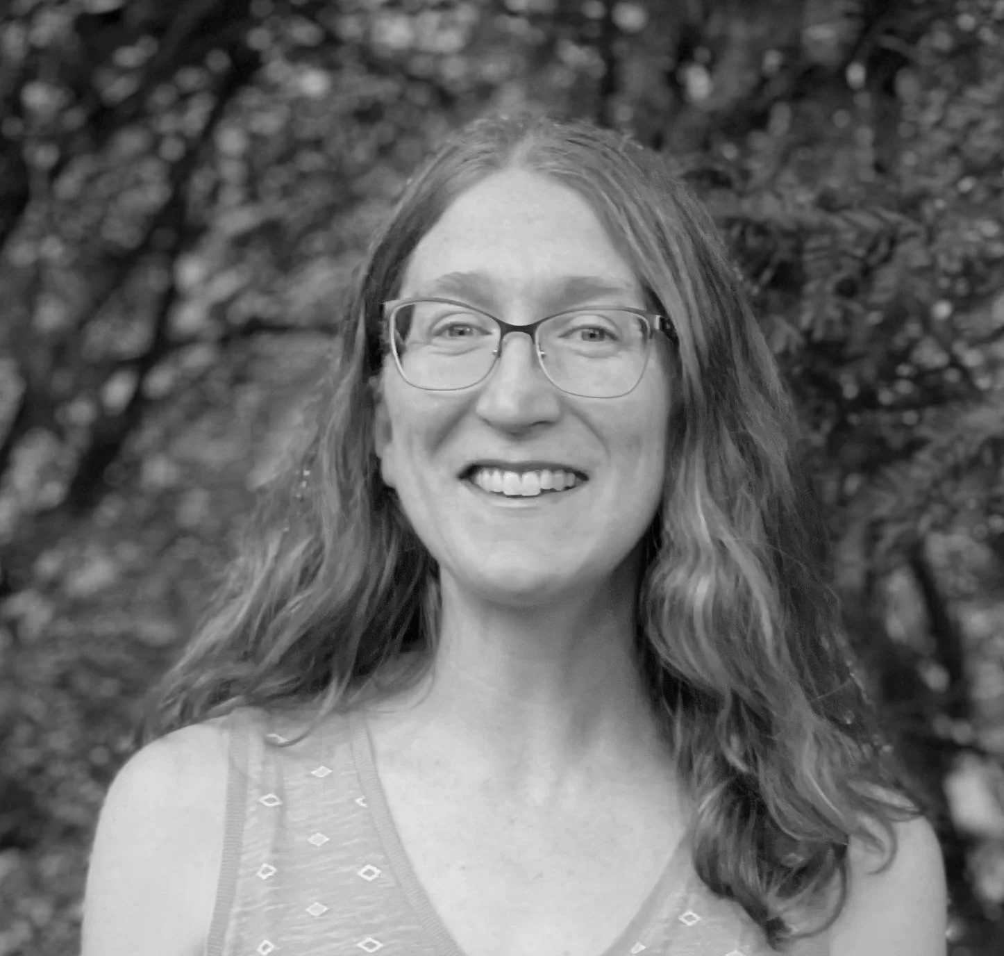 Black and white photo of a woman with glasses, long wavy hair, smiling, outdoors with trees in the background.