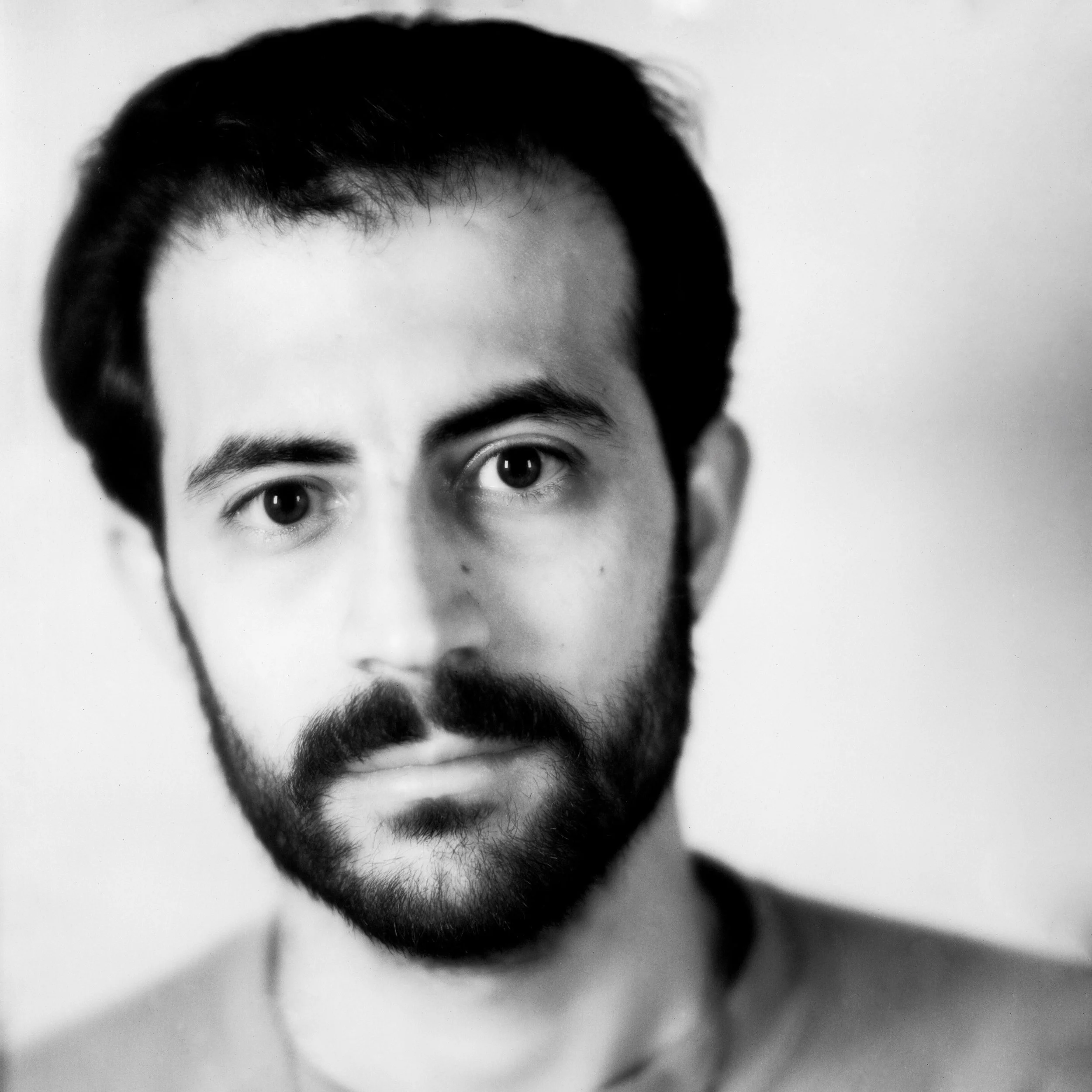 Black and white close-up portrait of a man with dark hair, beard, and mustache, looking directly at the camera with a serious expression.