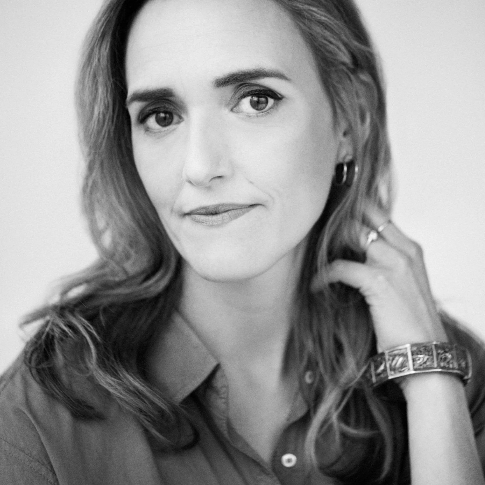 A black-and-white portrait of a woman with wavy hair, looking at the camera, wearing a buttoned shirt, earrings, and a bracelet.