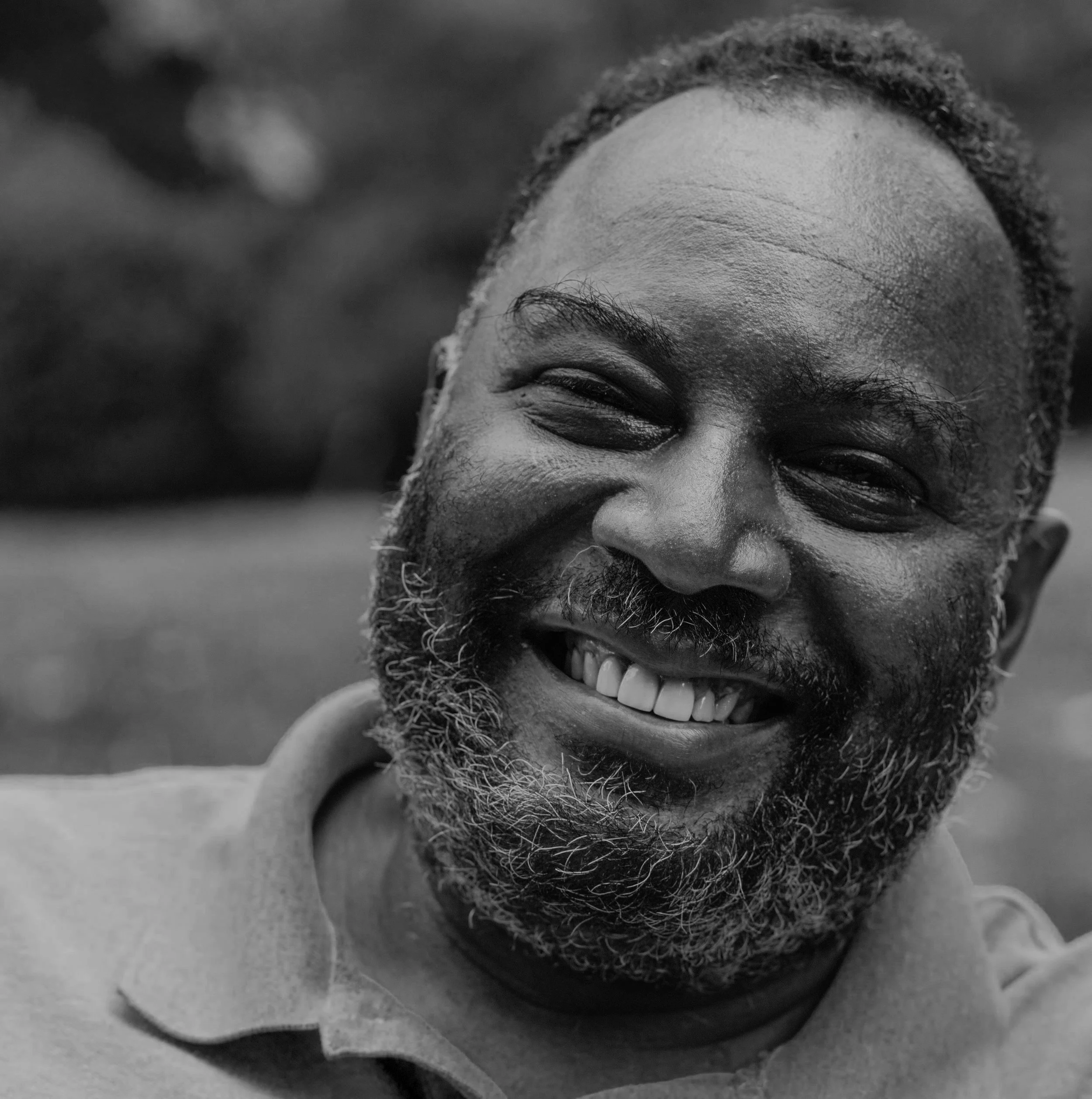 Close-up of a smiling man with a beard, showing teeth, outdoors, in black and white.