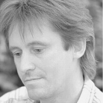 A black and white portrait of a man with medium-length, layered hair, wearing a checkered shirt, looking downward.