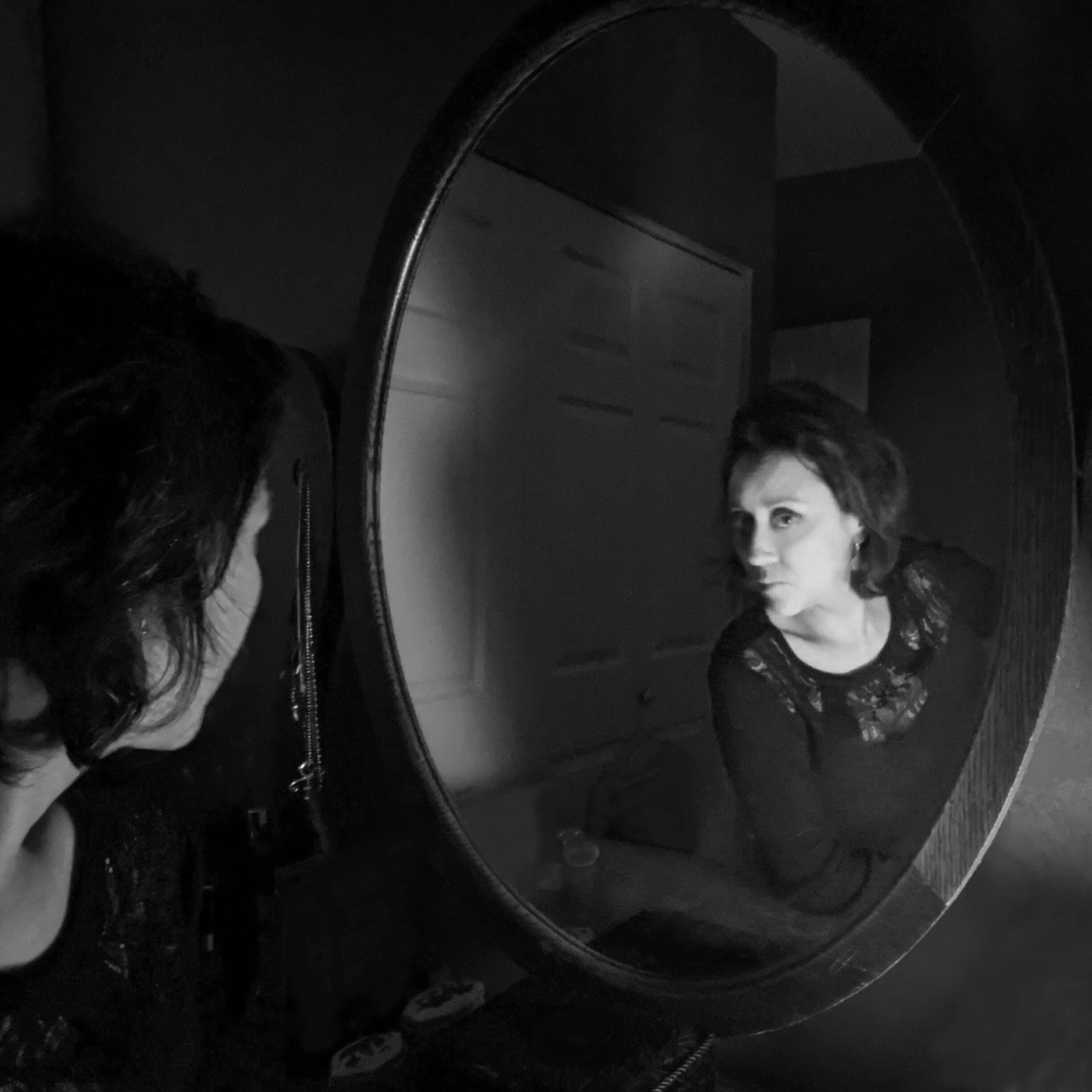 A woman with dark, wavy hair looking into a mirror, which reflects her face as she looks at herself with a serious expression.