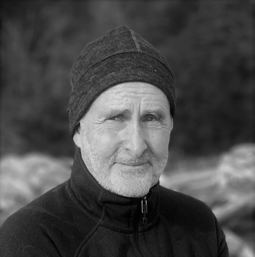 A black-and-white photo of an older man with a beard, wearing a beanie and a zip-up jacket outdoors.