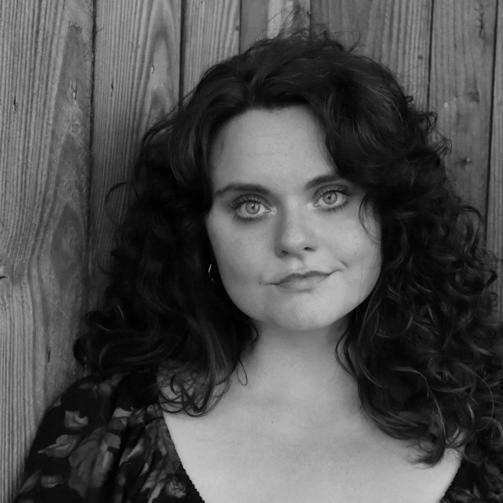 Black and white portrait of a woman with curly dark hair and light eyes, standing against a wooden wall.