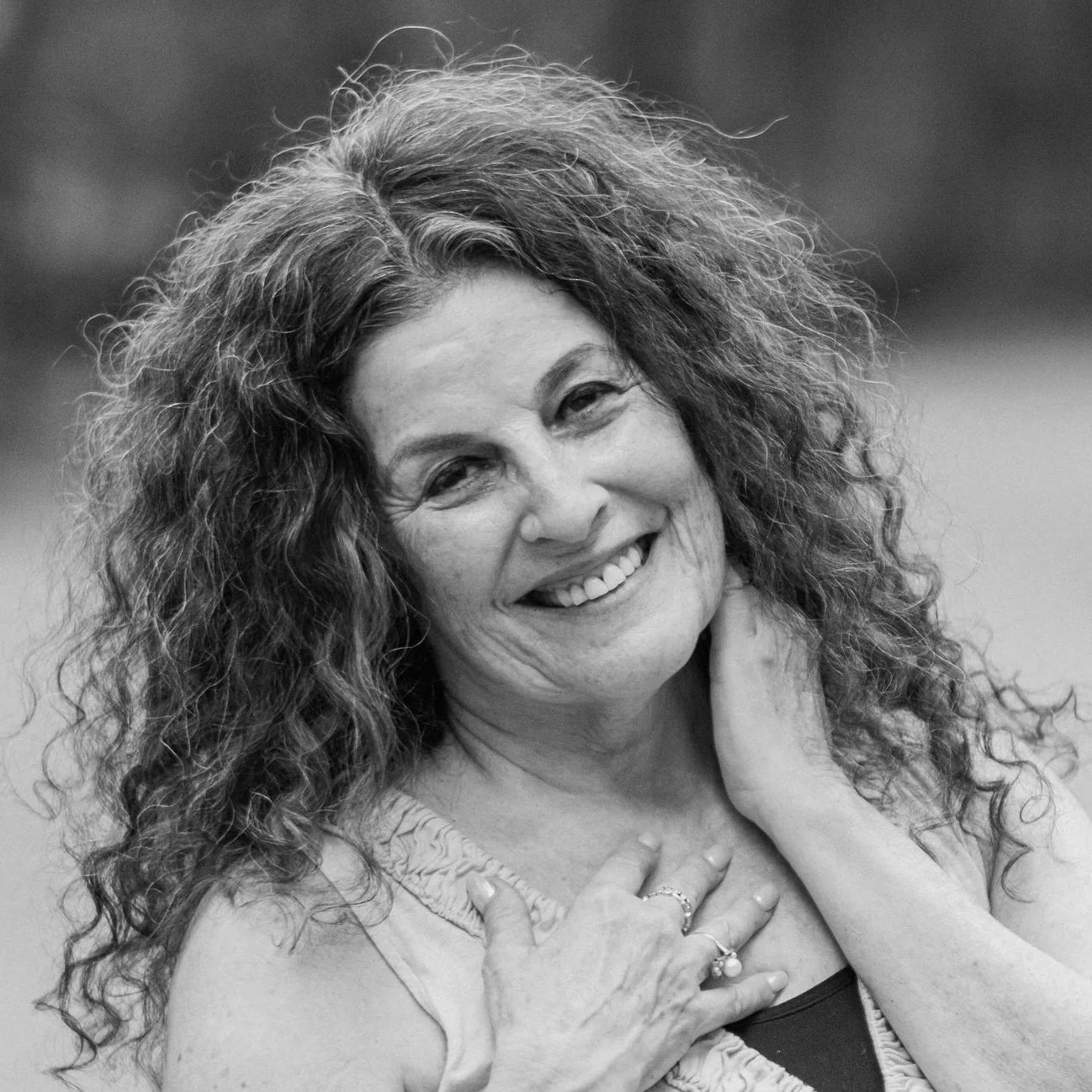 Black and white photo of a smiling woman with curly hair, touching her neck and chest with her hands.