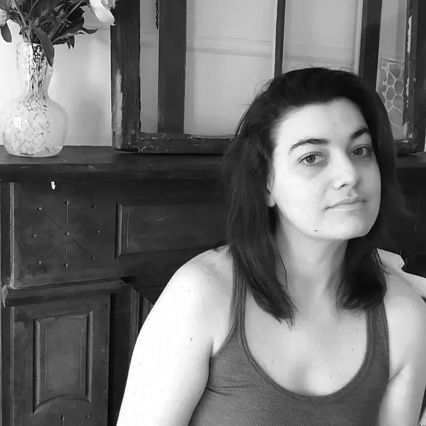 Black-and-white photo of a woman with shoulder-length dark hair, wearing a tank top, sitting indoors in front of a wooden cabinet with a flower vase on top.