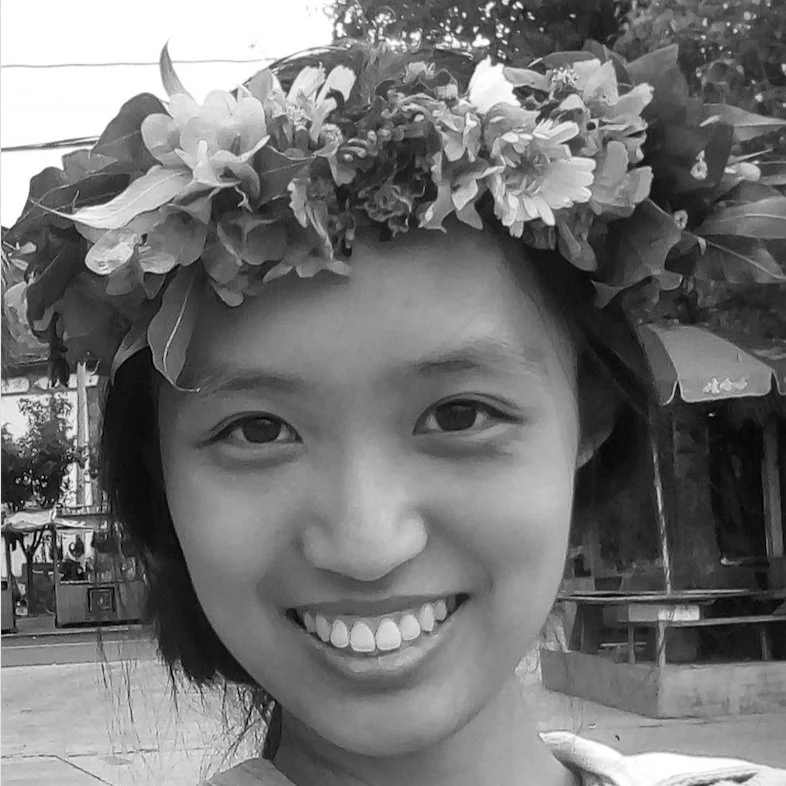 Young woman smiling, wearing a floral crown, outdoors in a park or fairground setting.
