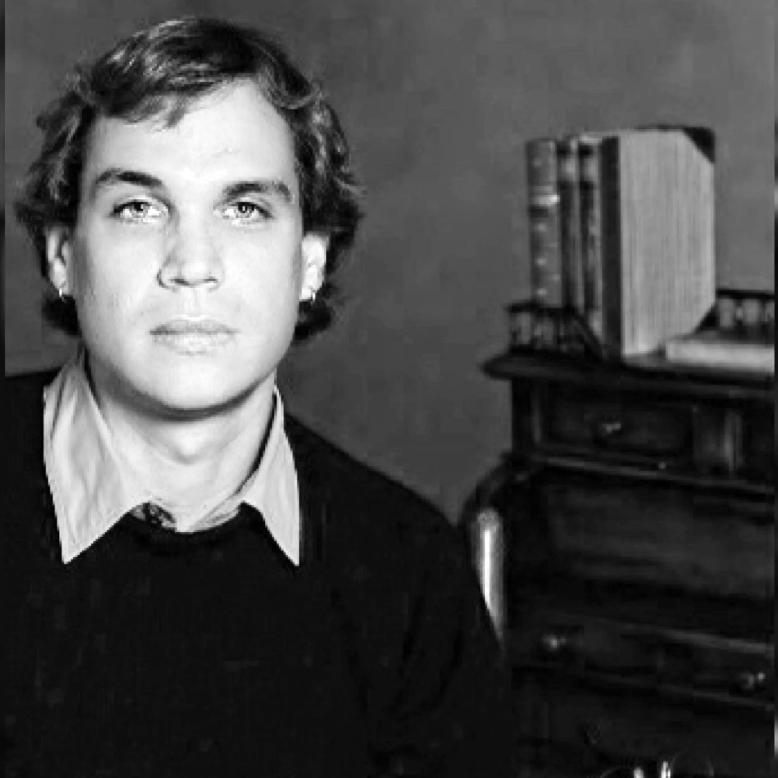 Black and white photo of a woman with short hair, wearing a collared shirt and dark sweater, sitting in front of a desk with books on it.
