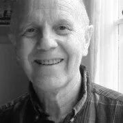 Smiling man with short hair wearing a checkered shirt, standing indoors near a window.