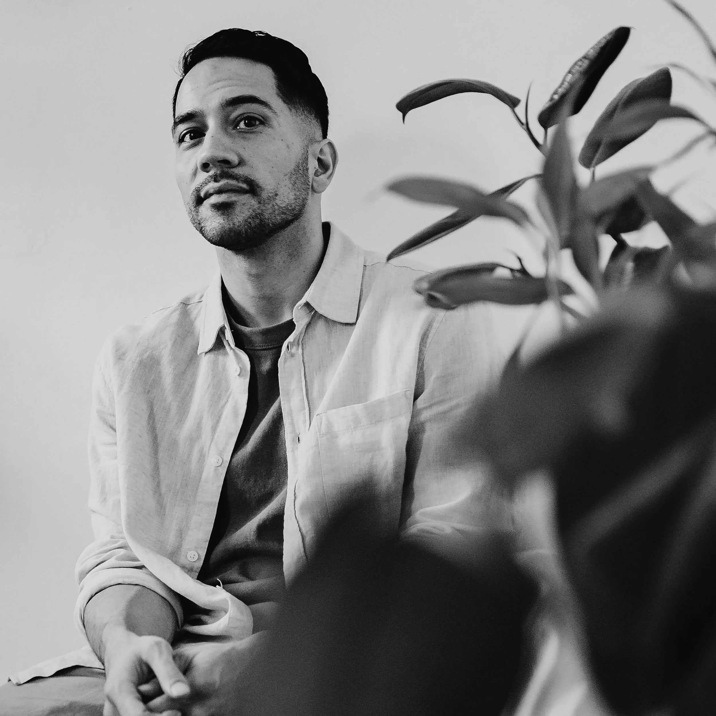 Black and white photo of a man with short dark hair and a beard, wearing a light-colored shirt, sitting indoors with a leafed plant in the foreground.