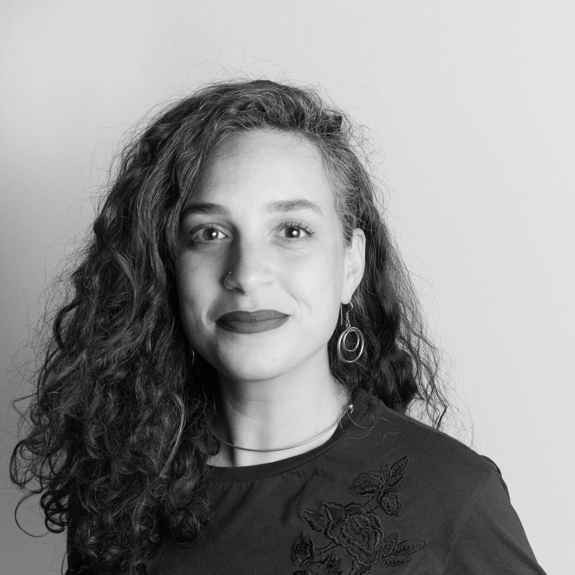 Black and white portrait of a woman with long, curly hair, wearing earrings, a necklace, and a dark top with floral embroidery on the shoulder, standing against a plain background.