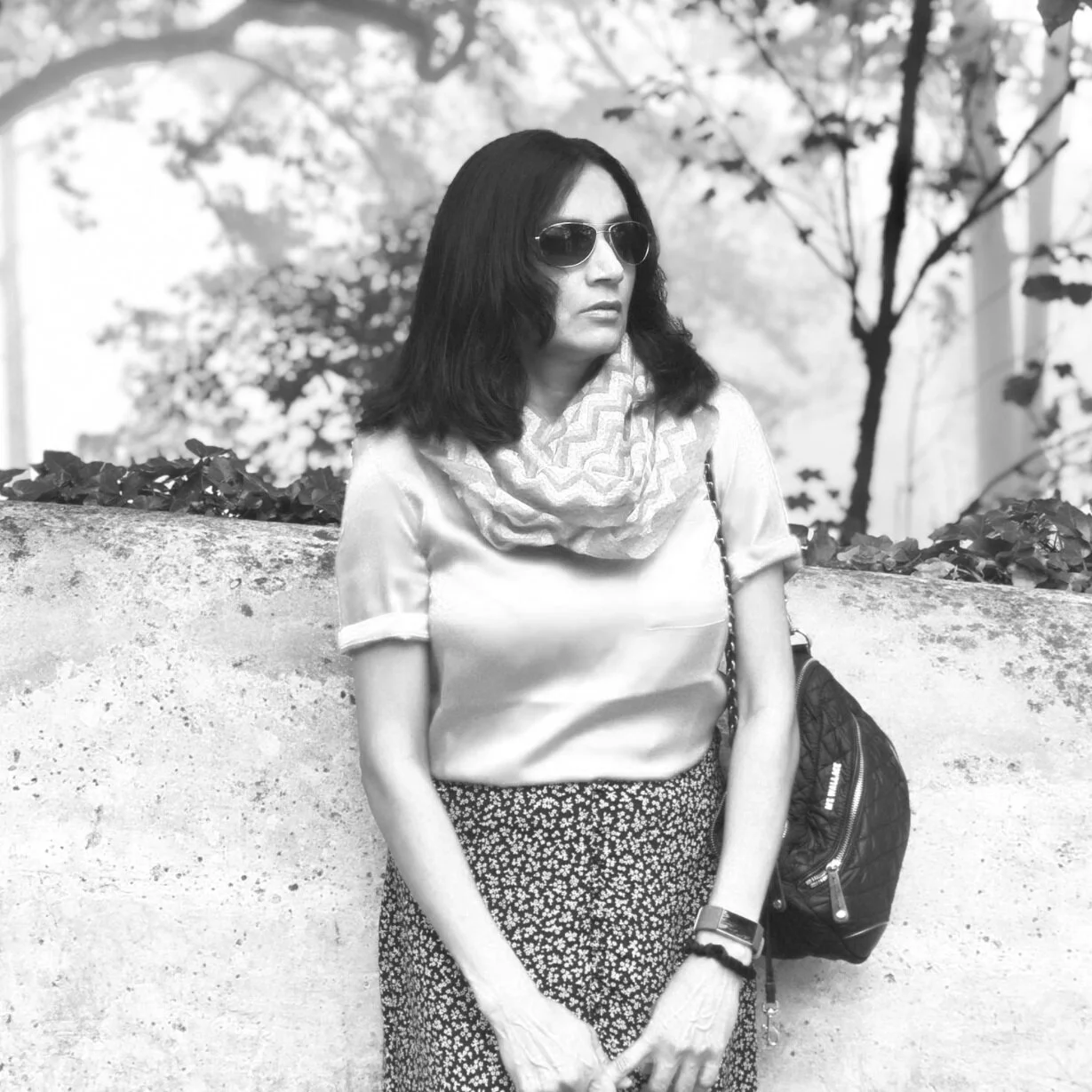 A woman with dark hair wearing sunglasses, a patterned scarf, a satin blouse, and a floral skirt, standing outdoors next to a concrete wall with trees and foliage in the background.