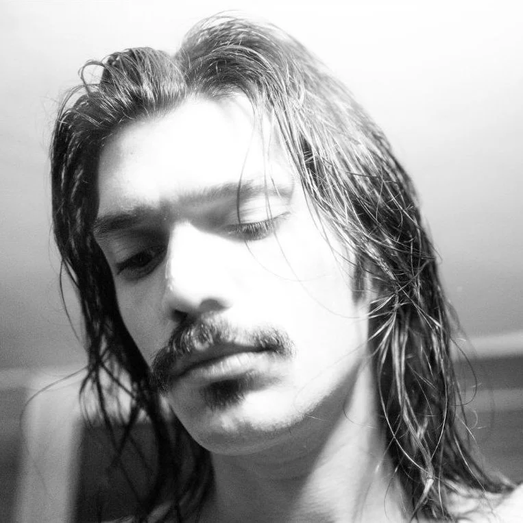 Black and white close-up photo of a young man with wet, long hair and a mustache, looking slightly down.