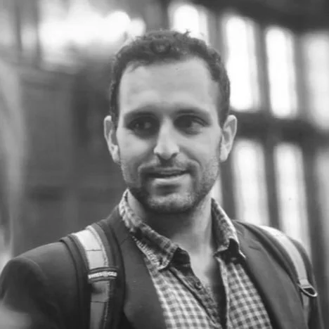 A black and white photo of a man with short curly hair and a beard, wearing a checkered shirt and backpack, standing indoors near large windows.