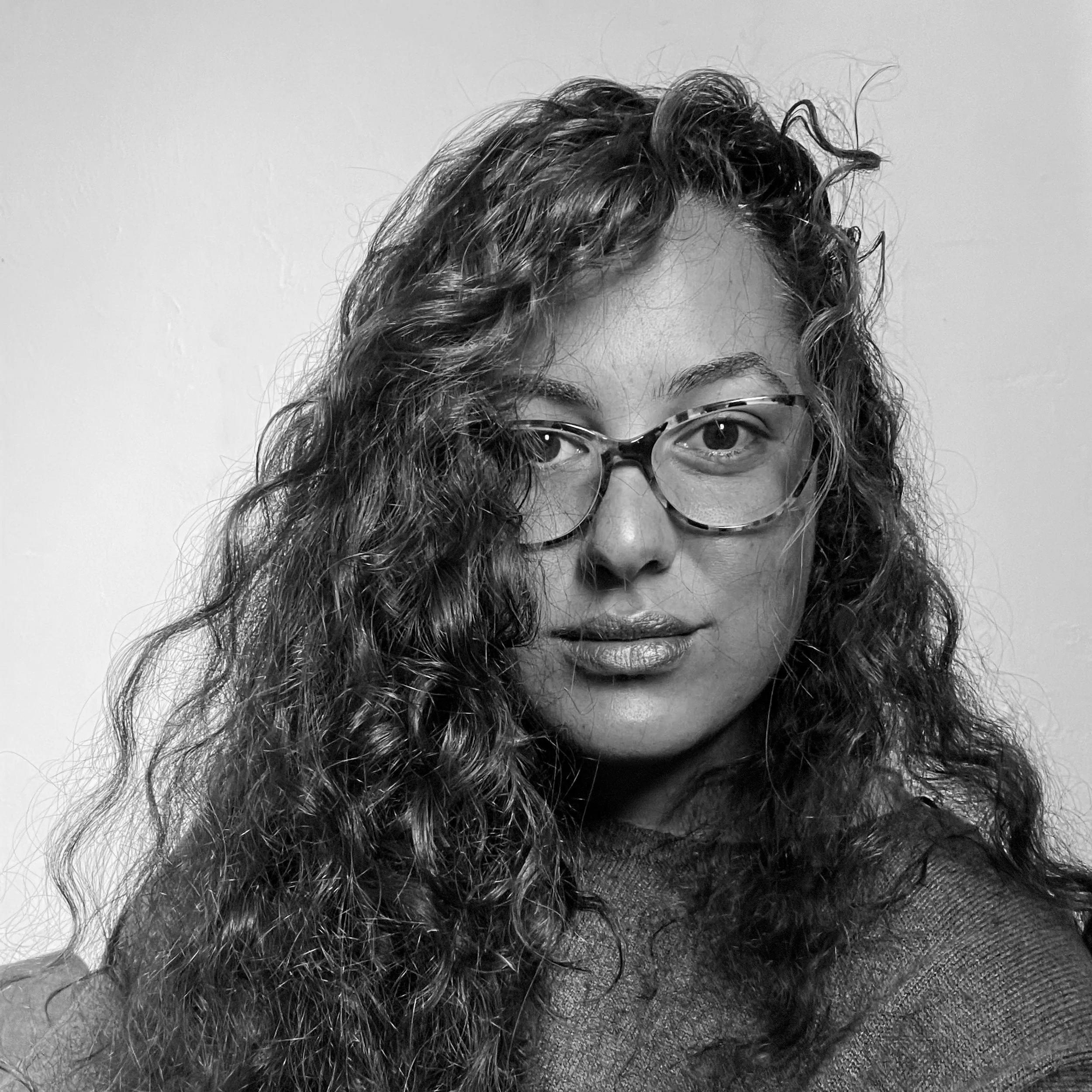 A woman with curly hair wearing glasses, looking directly at the camera against a plain background.
