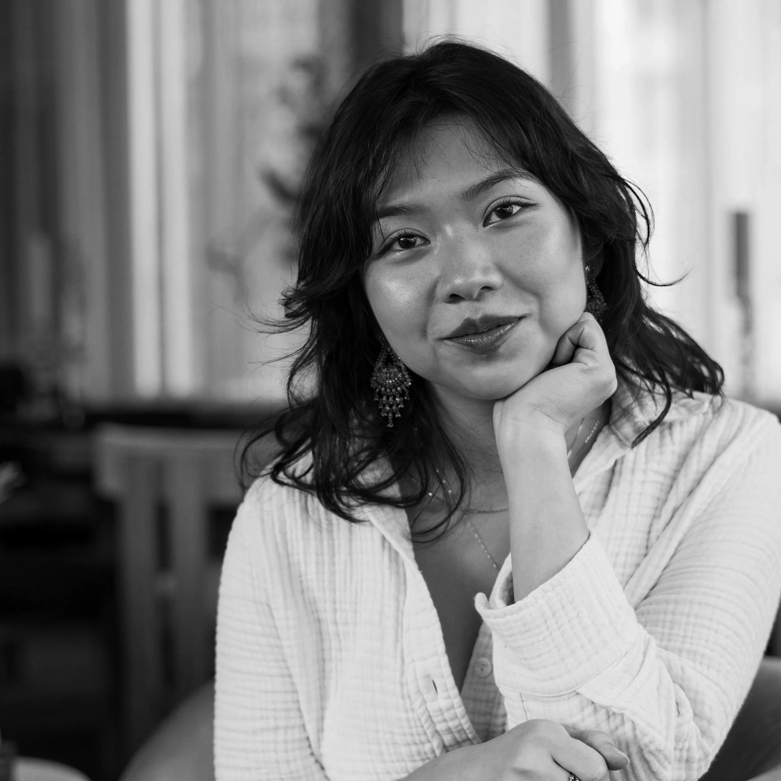 A black and white close-up portrait of an Asian woman with shoulder-length wavy hair, wearing earrings and a light-colored shirt, resting her chin on her hand and smiling softly.