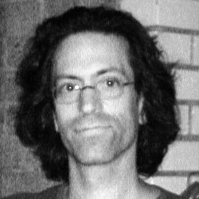 Black and white photo of a man with shoulder-length curly hair wearing glasses, against a brick wall background.